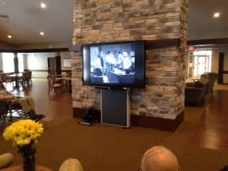 Interior view of a common area in an assisted living facility with a stone pillar holding a flat-screen TV playing a black and white show. There are tables and chairs in the background, a vase with yellow flowers in the foreground, and two people partially visible sitting and watching the TV.