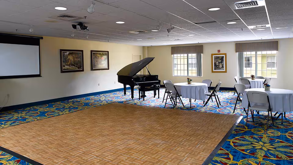 A spacious room with a patterned blue carpet and a wooden dance floor area in the center. There is a black grand piano near the wall, round tables covered with white tablecloths, and folding chairs arranged around the tables. The walls are light-colored with framed artwork, and there are windows letting in natural light.