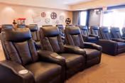 A row of six black leather recliner chairs with cup holders in a cozy room with beige carpet and walls. The back wall is decorated with vintage film reels and movie-themed decor, suggesting a small theater or media room setting.