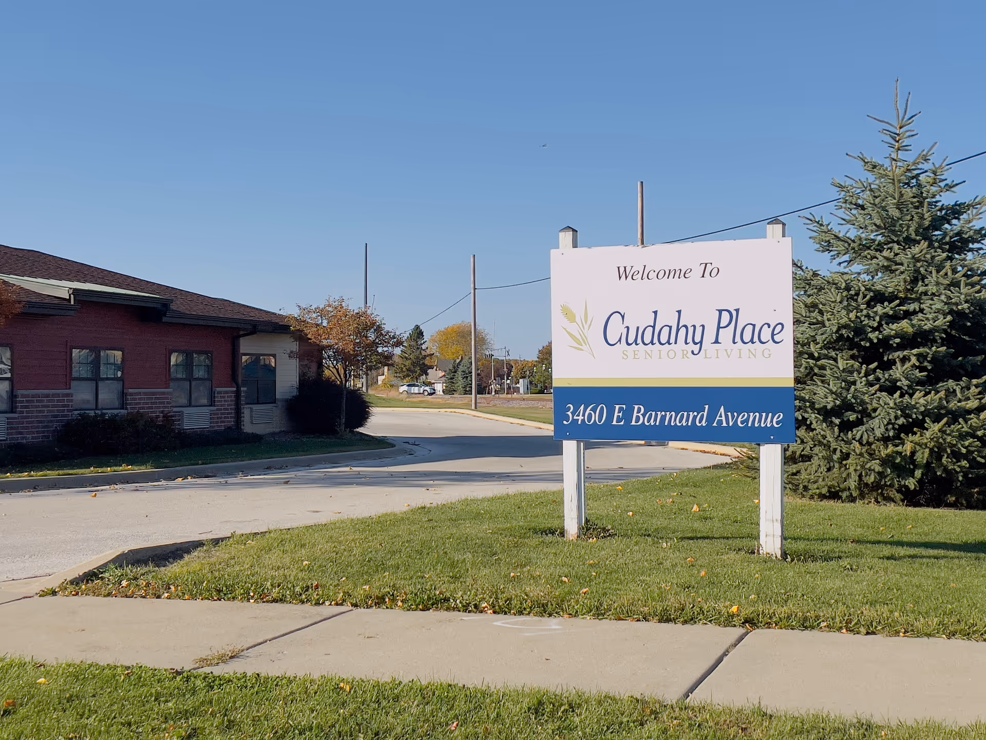Outdoor view of the entrance area of Cudahy Place Senior Living, featuring a large white sign with the facility name and address 3460 E Barnard Avenue, a red brick building on the left, green grass, a sidewalk, and a clear blue sky.