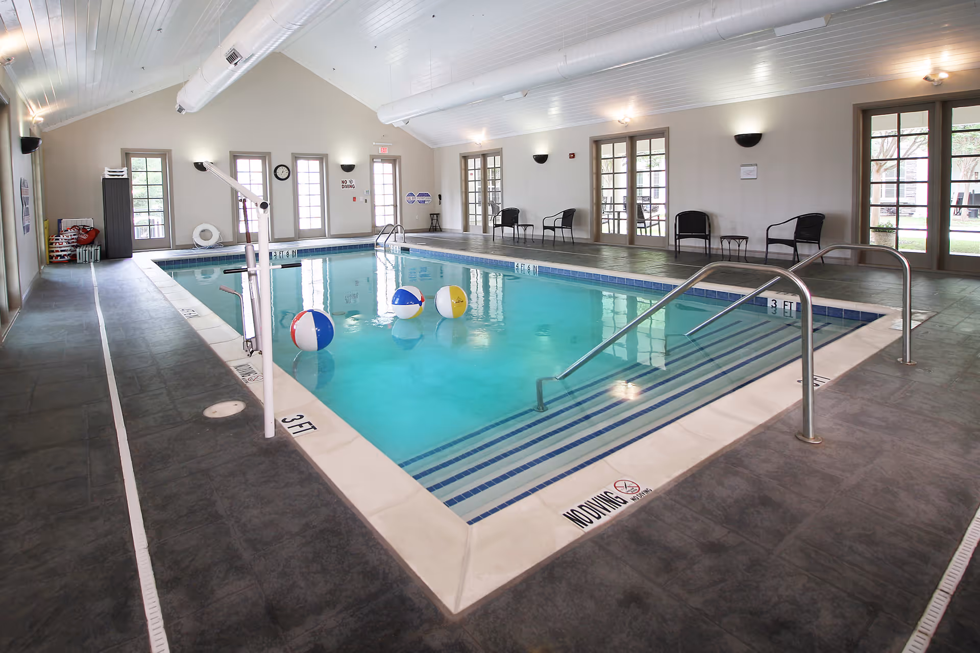 Indoor swimming pool with clear blue water, three colorful beach balls floating on the surface, metal handrails for pool entry, and several black chairs along the walls. The room has a high white ceiling with exposed beams and multiple glass doors letting in natural light.