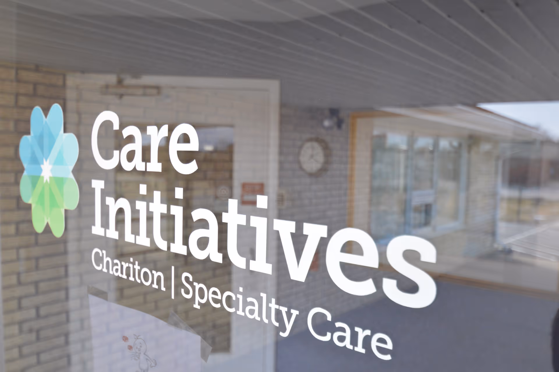 Glass door with the logo and text 'Care Initiatives Chariton Specialty Care' printed on it, reflecting the interior of a building with brick walls, a clock, and windows.