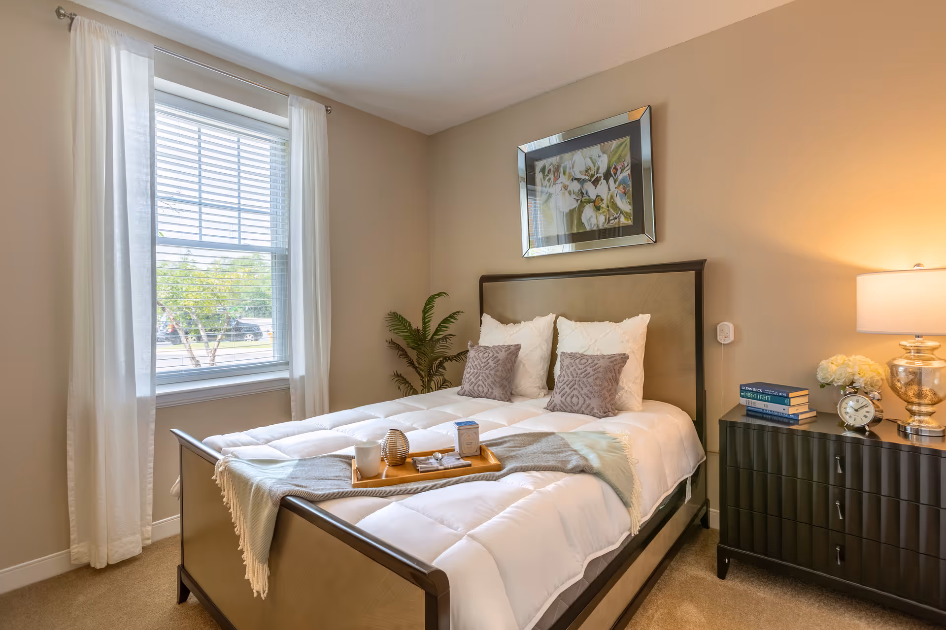 Well-lit bedroom with a made bed, decorative pillows, a nightstand with a lamp and books, a window with curtains, and framed artwork above the headboard.