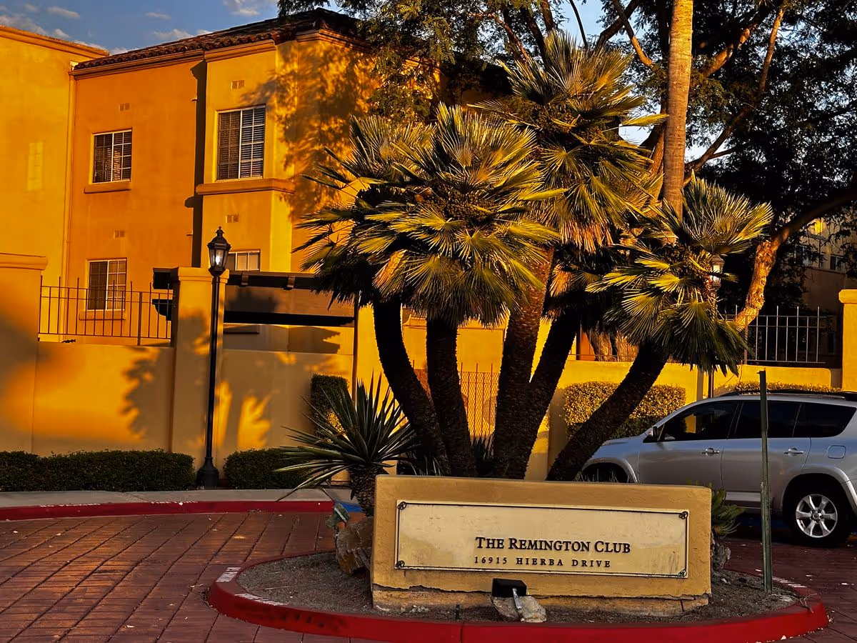 Entrance sign reading 'The Remington Club' in front of palm trees, a parked car, and the club building bathed in warm sunlight.