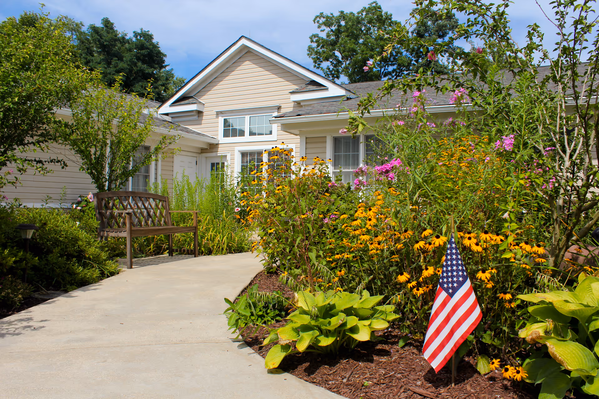 Front entrance of a light-colored assisted living building with a walkway, bench, colorful flower beds and an American flag.
