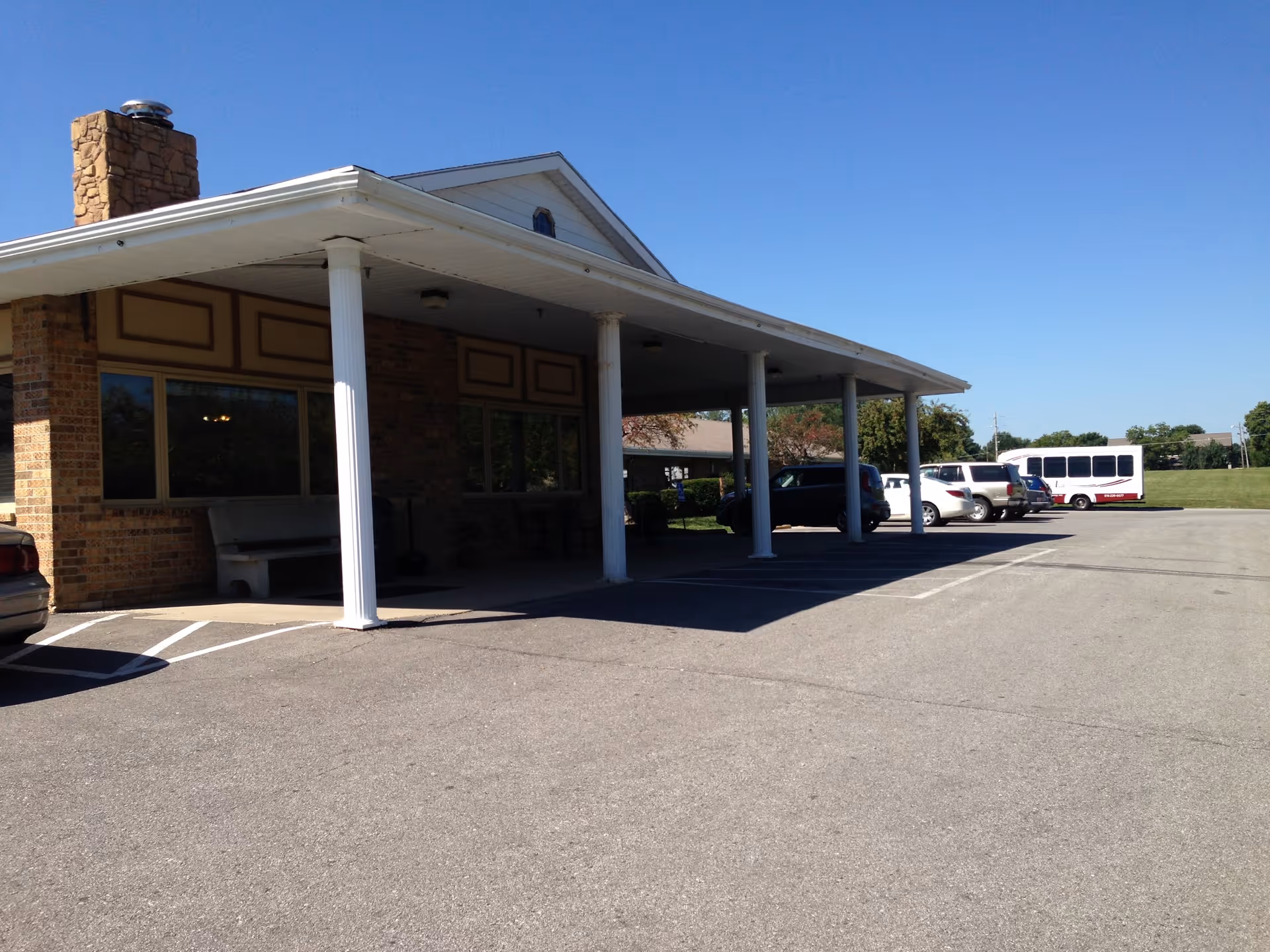 Exterior view of Shangri-La Rehab & Living Center showing a covered entrance supported by white columns, a brick chimney, several parked cars, and a shuttle bus in the parking lot under a clear blue sky.