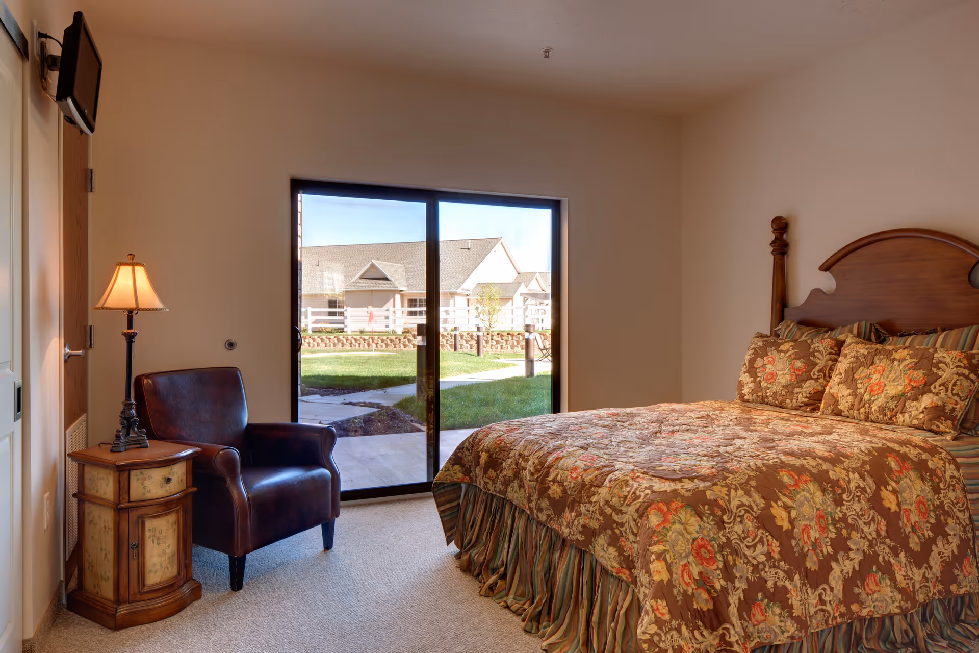 A furnished bedroom with a floral-patterned bed, leather armchair, side table and sliding glass door opening to a courtyard.