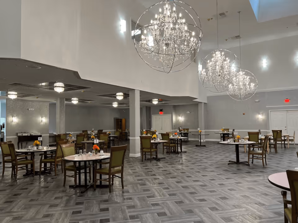 A spacious dining room with multiple round tables and chairs arranged neatly. Each table has a small floral centerpiece and place settings. The room features elegant chandeliers with crystal details hanging from the ceiling, gray walls, and a patterned gray floor.
