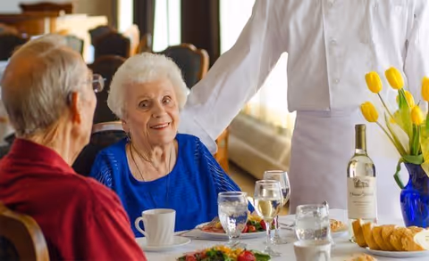 An elderly woman in a blue top and an elderly man in a red shirt are seated at a dining table with plates of food, glasses of water, and a bottle of wine. A waiter dressed in white is standing beside them, serving or attending to the table. Yellow tulips in a blue vase are also on the table.