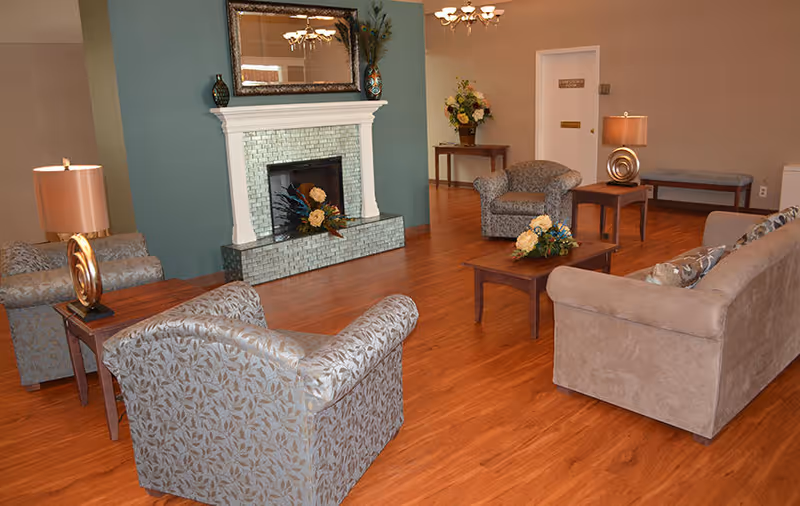 A cozy living room area with wooden flooring, featuring a beige sofa, two patterned armchairs, a wooden coffee table with floral arrangements, a white fireplace with decorative vases on the mantel, and two side tables with lamps. In the background, there is a small table with a flower arrangement and a bench against the wall.