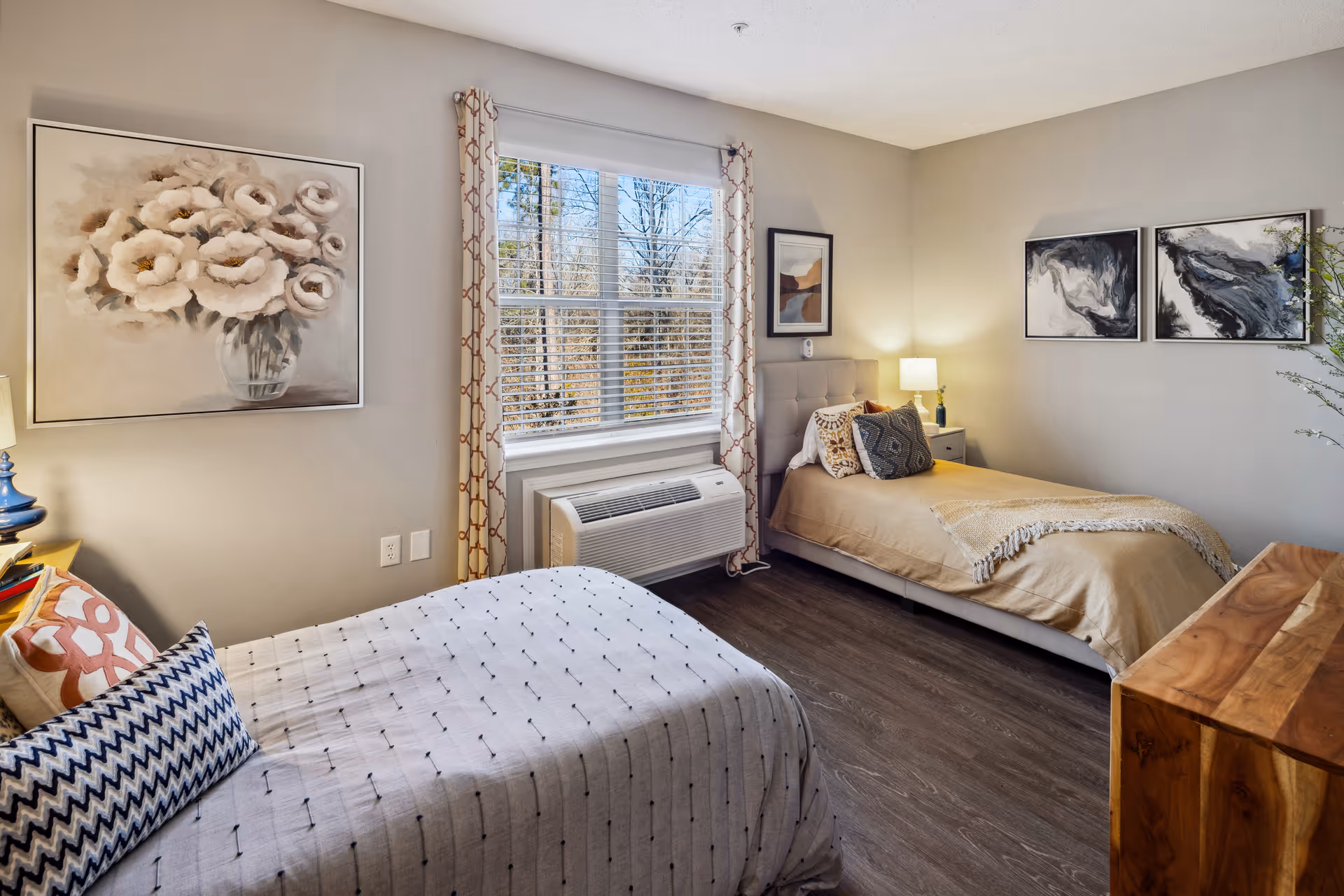 A cozy bedroom with two single beds, each with decorative pillows and blankets. There is a window with patterned curtains and an air conditioning unit below it. The walls are adorned with framed artwork, including a large painting of white flowers and two abstract black and white paintings. A wooden dresser is visible on the right side of the room, and a bedside table with a lamp is placed between the beds.