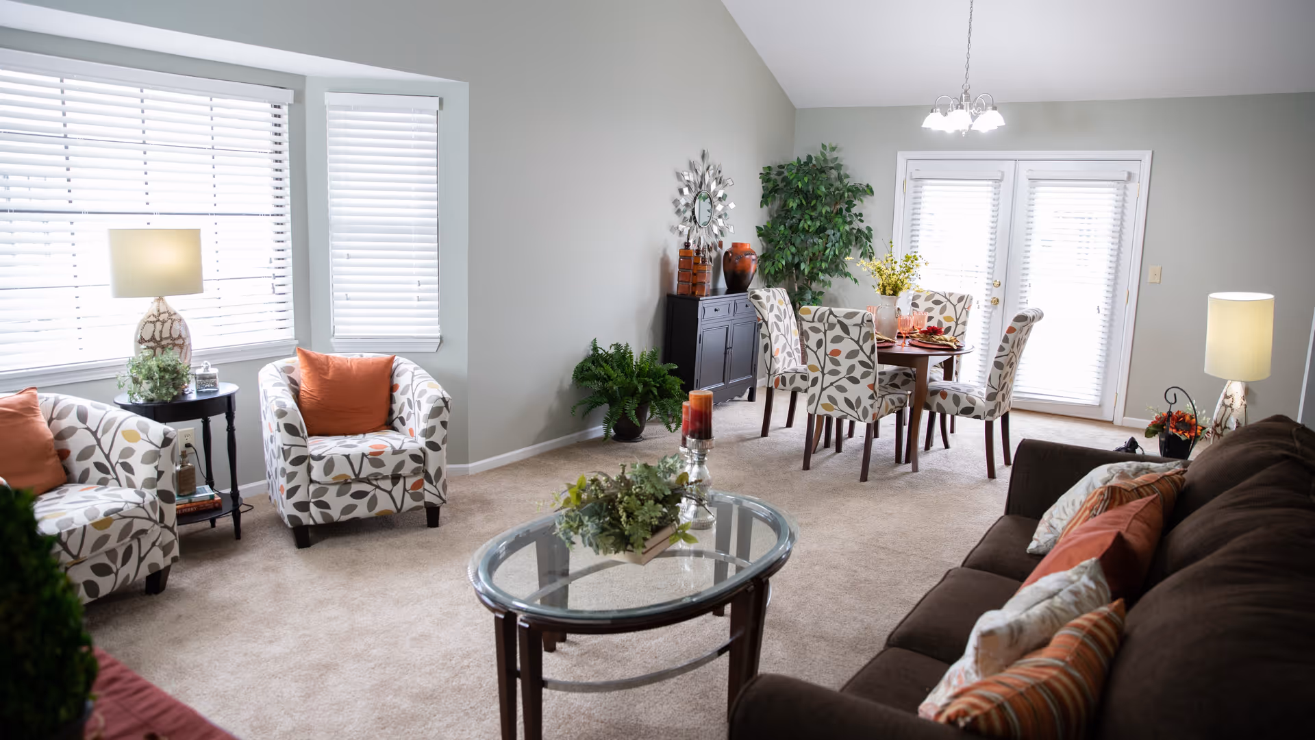 A bright and cozy living and dining area with beige carpet, a dark brown sofa with multiple cushions, two patterned armchairs with orange pillows, a glass coffee table with decorative greenery, a round dining table with four patterned chairs, and large windows with white blinds letting in natural light. There are also two table lamps, a black sideboard with decorative items, and several green plants.