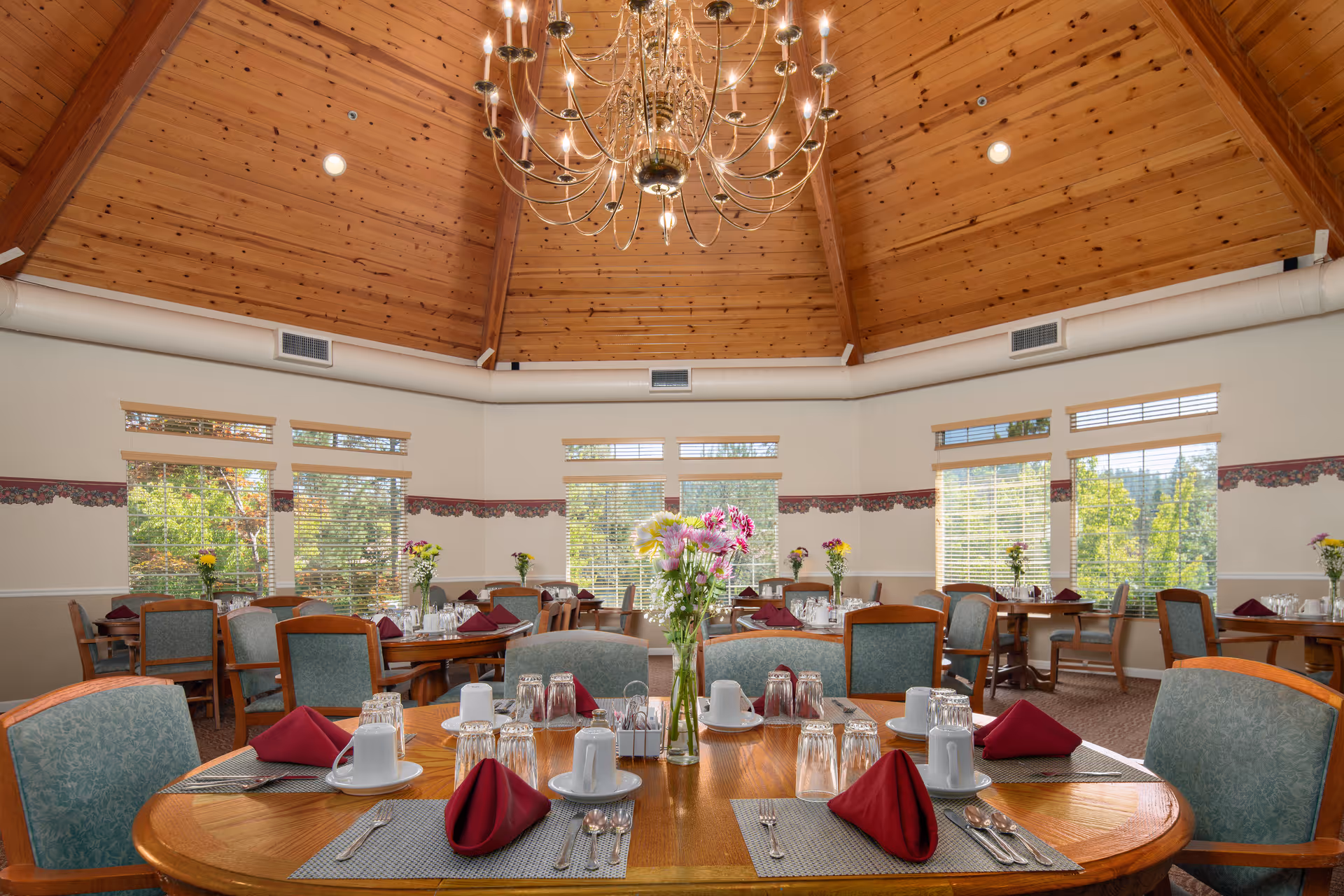 Dining room with round wooden tables set with white cups, glasses, silverware, and burgundy napkins. The room has large windows with blinds, a high wooden ceiling with a chandelier, and floral wallpaper border. Several tables have flower vases with colorful flowers.