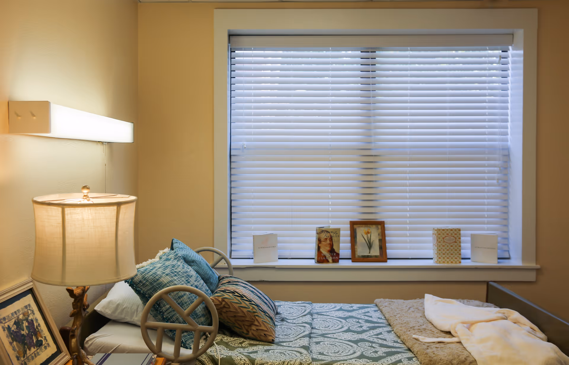A cozy bedroom with a single bed featuring patterned bedding and multiple pillows. A lamp with a beige shade sits on a bedside table next to the bed. The window behind the bed has white blinds and a windowsill decorated with framed pictures and cards.