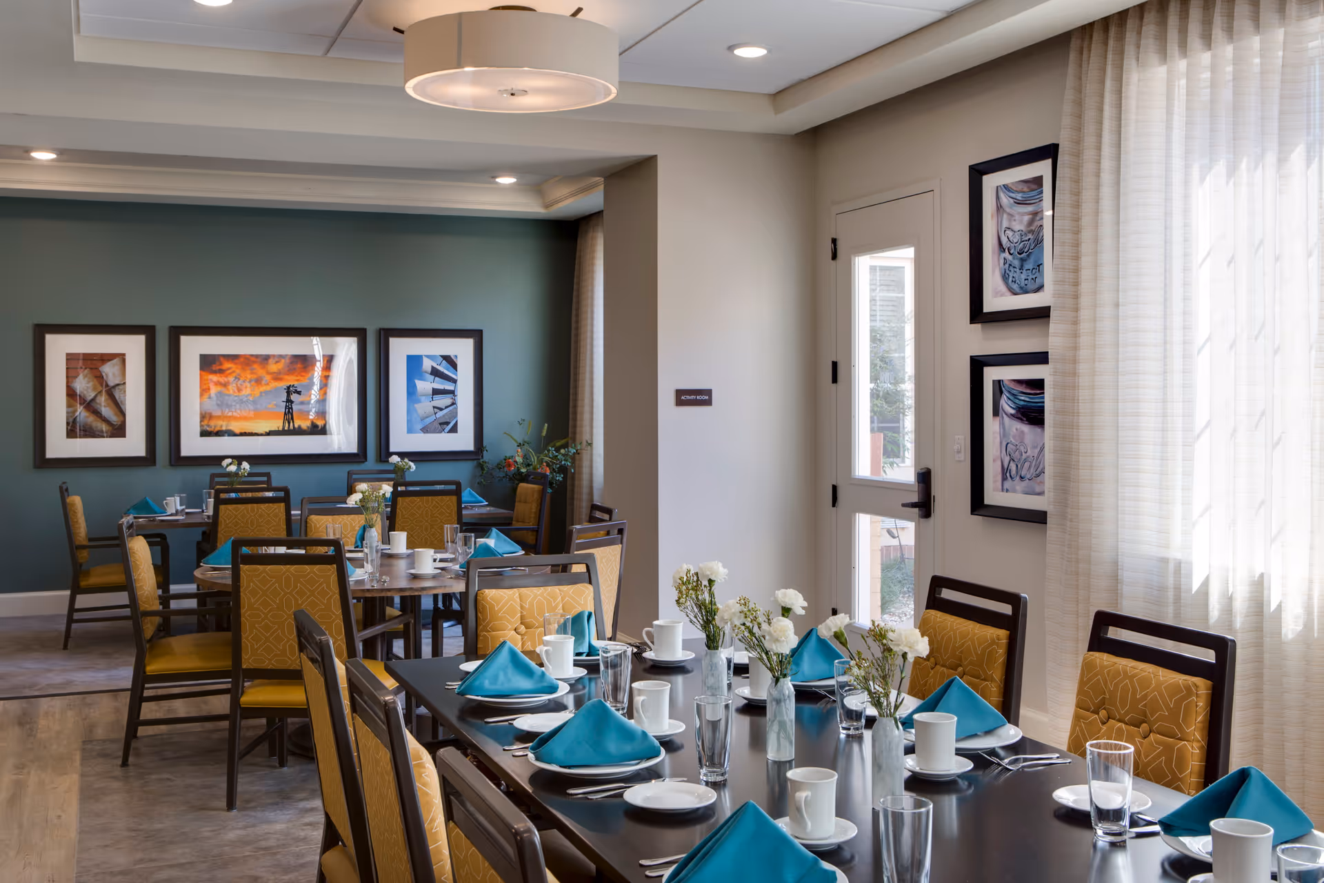 A dining room in The Legacy at South Plains featuring multiple tables set with white plates, cups, glasses, silverware, and teal napkins. The chairs have mustard yellow upholstery with a geometric pattern. The room has light-colored walls, a teal accent wall with three framed pictures, and a door with glass panels. White curtains cover the windows, and small vases with white flowers are placed on the tables.