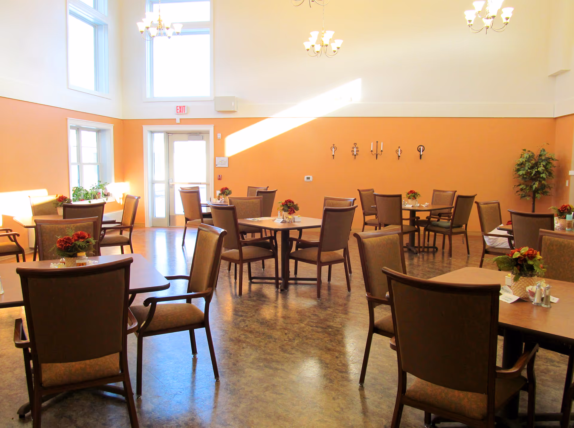 A bright dining room with multiple tables and chairs arranged neatly. Each table has a small floral centerpiece. The walls are painted orange on the lower half and white on the upper half. Large windows and glass doors allow natural light to fill the room. There are chandeliers hanging from the ceiling and decorative wall sconces on the orange wall.