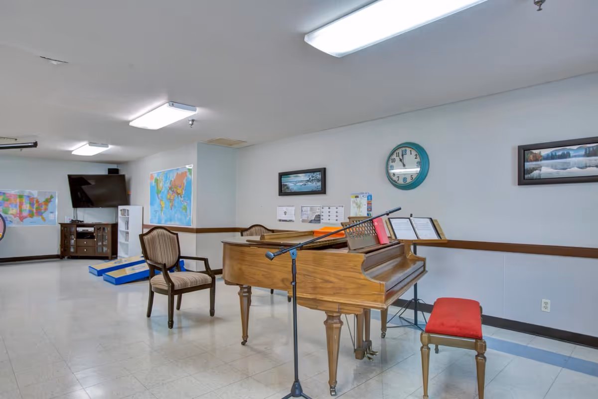 A spacious room with a grand piano and a red cushioned piano bench. There is a microphone stand in front of the piano and a wooden chair nearby. The walls are decorated with framed landscape pictures and a round clock. In the background, there is a TV mounted on the wall, a cabinet, a bookshelf, and two large maps on the walls. The floor is tiled and the ceiling has fluorescent lights.
