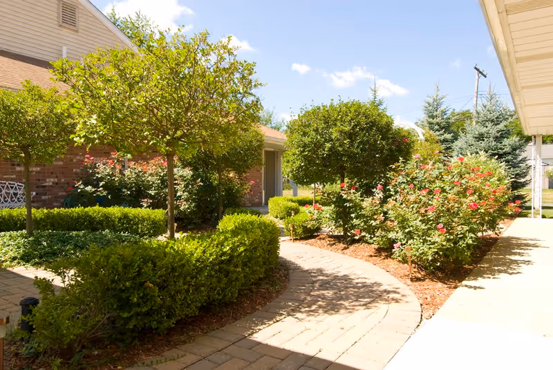 A sunny outdoor garden area at Villa Marie featuring a paved walkway winding through neatly trimmed bushes, small trees, and flowering plants. The garden is adjacent to a brick building and a covered walkway.
