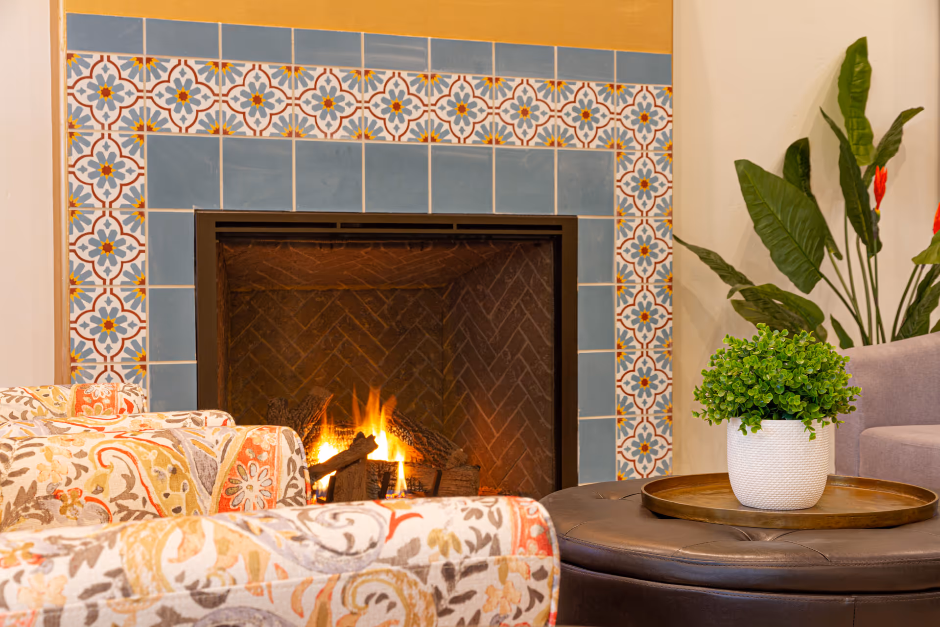 A cozy living room area featuring a lit fireplace surrounded by decorative blue and patterned tiles. In front of the fireplace are two upholstered chairs with a colorful floral pattern. A round leather ottoman holds a wooden tray with a white pot containing a green leafy plant. In the background, there is a tall green plant with red flowers against a beige wall.