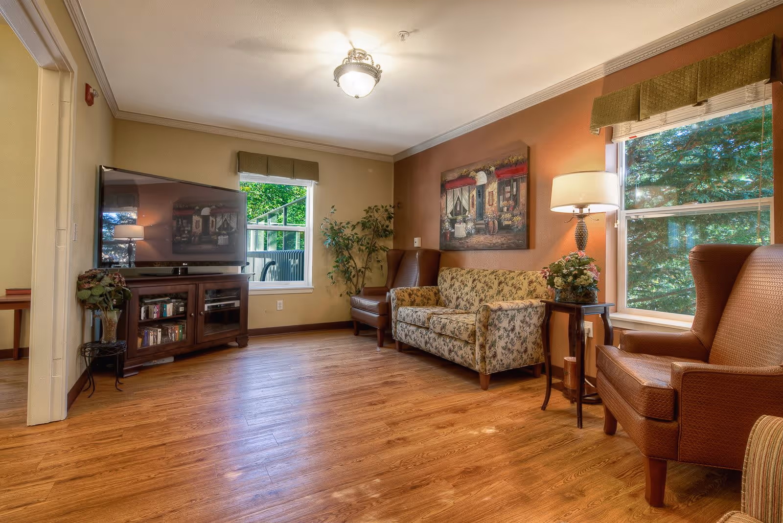 A cozy living room with wooden flooring, a floral patterned sofa, two brown armchairs, a wooden TV stand with a flat-screen TV, a side table with a lamp and a flower arrangement, two windows with green valances showing trees outside, and a painting on the wall.