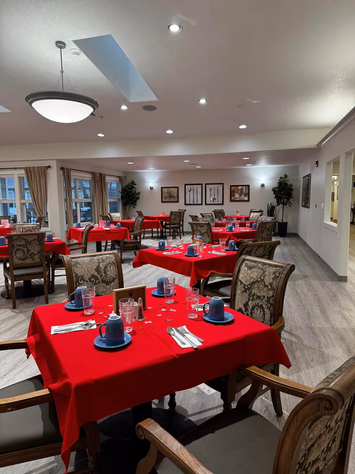 A dining room with multiple tables covered in red tablecloths, each set with blue cups, plates, glasses, and silverware. The room has patterned upholstered chairs, large windows with beige curtains, framed artwork on the walls, and potted plants in the corners. The ceiling has recessed lighting and a hanging light fixture.