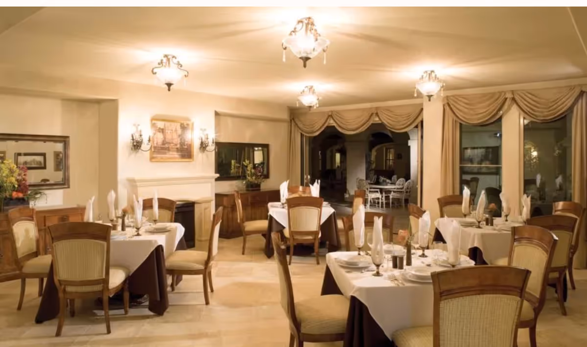 Elegant dining room with several tables covered in white tablecloths and brown table skirts, each set with plates, glasses, and neatly folded napkins. The room features wooden chairs with cushioned seats, warm lighting from ceiling fixtures and wall sconces, a fireplace, and large windows with draped curtains. In the background, there is an adjoining outdoor seating area with white chairs and tables.