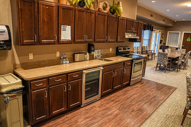 Interior view of a senior living facility kitchen area with wooden cabinets, a countertop with a sink, a stove, a small refrigerator, and a trash bin. In the background, there is a dining area with tables and chairs, and large windows letting in natural light.