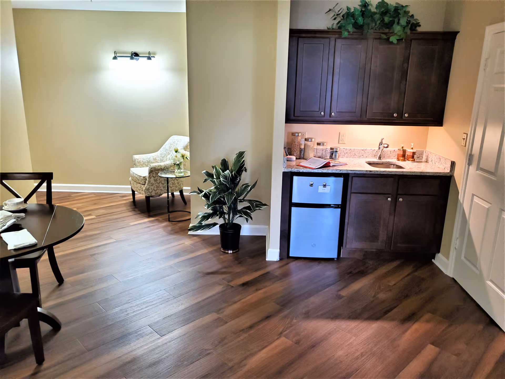 Interior view of a senior living facility room at Madison at Ocoee featuring a small kitchenette with dark wood cabinets, a granite countertop, a mini refrigerator, and a sink. To the left, there is a cozy sitting area with a patterned armchair and a small round glass table with a flower vase. A wooden dining table with chairs is partially visible in the foreground. The room has wood flooring and beige walls with a wall-mounted light fixture.