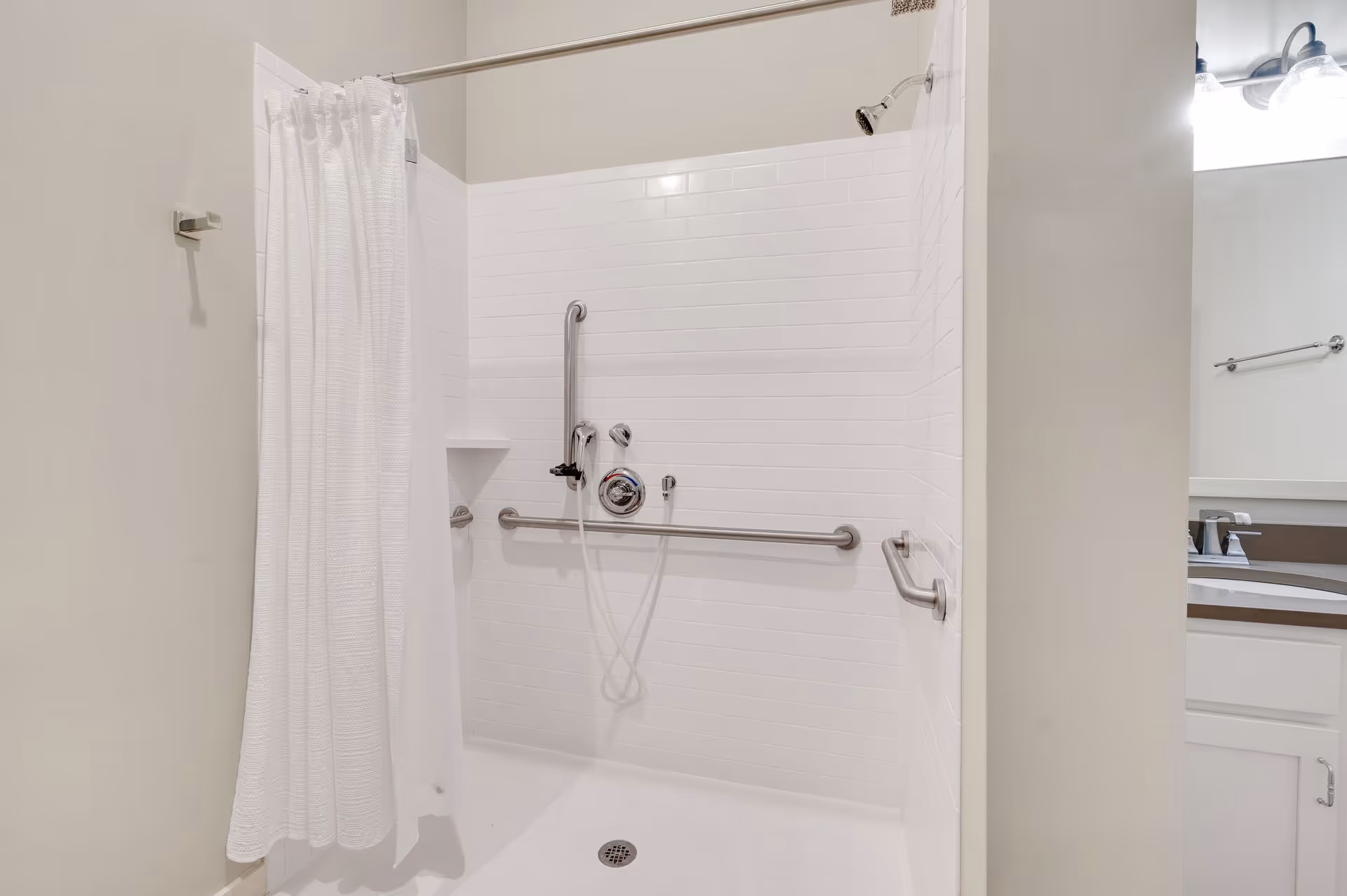 A clean, accessible shower area with white tiled walls, a white shower curtain, and multiple stainless steel grab bars installed for safety. The shower has a handheld showerhead and a small built-in shelf. Part of a bathroom sink and mirror are visible to the right.