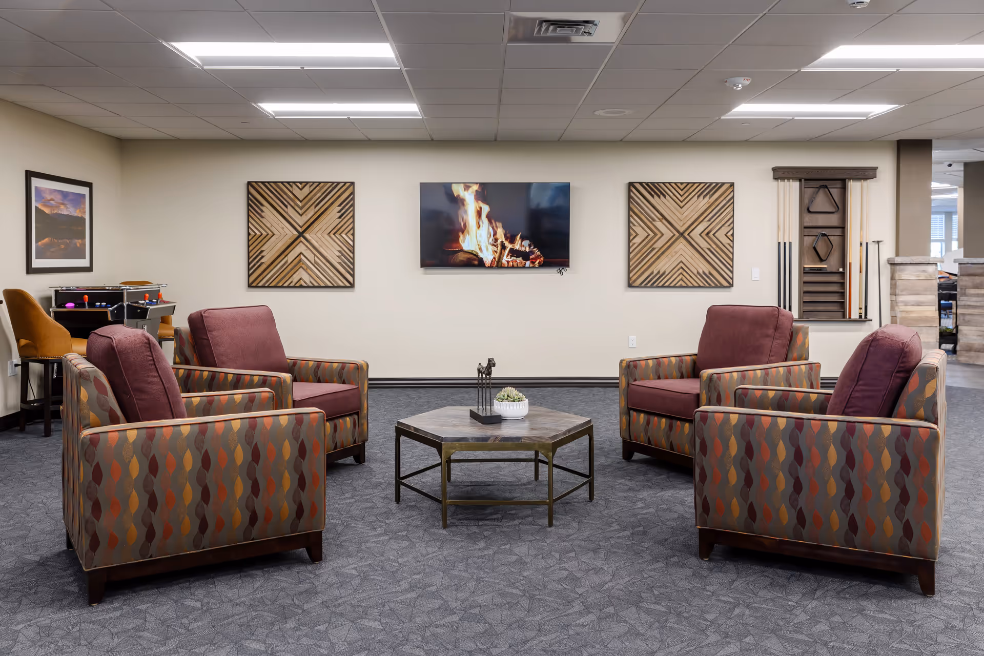 A cozy senior living common area with four patterned armchairs arranged around a hexagonal wooden coffee table. A flat-screen TV on the wall displays a burning fireplace. The walls are decorated with two geometric wooden art pieces and a framed landscape photograph. In the background, there is a small foosball table with two brown chairs and a wall-mounted rack holding pool cues and a triangle rack.