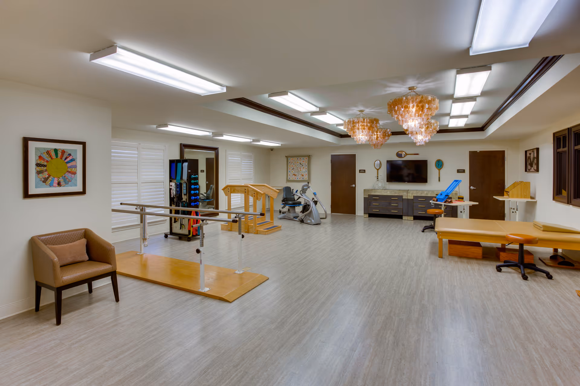 A spacious rehabilitation/therapy room with parallel walking bars, exercise machines, treatment tables, and seating.