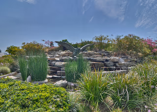 Landscaped outdoor garden with a tiered stone waterfall topped by a metal bird sculpture, surrounded by grasses and shrubs under a blue sky.