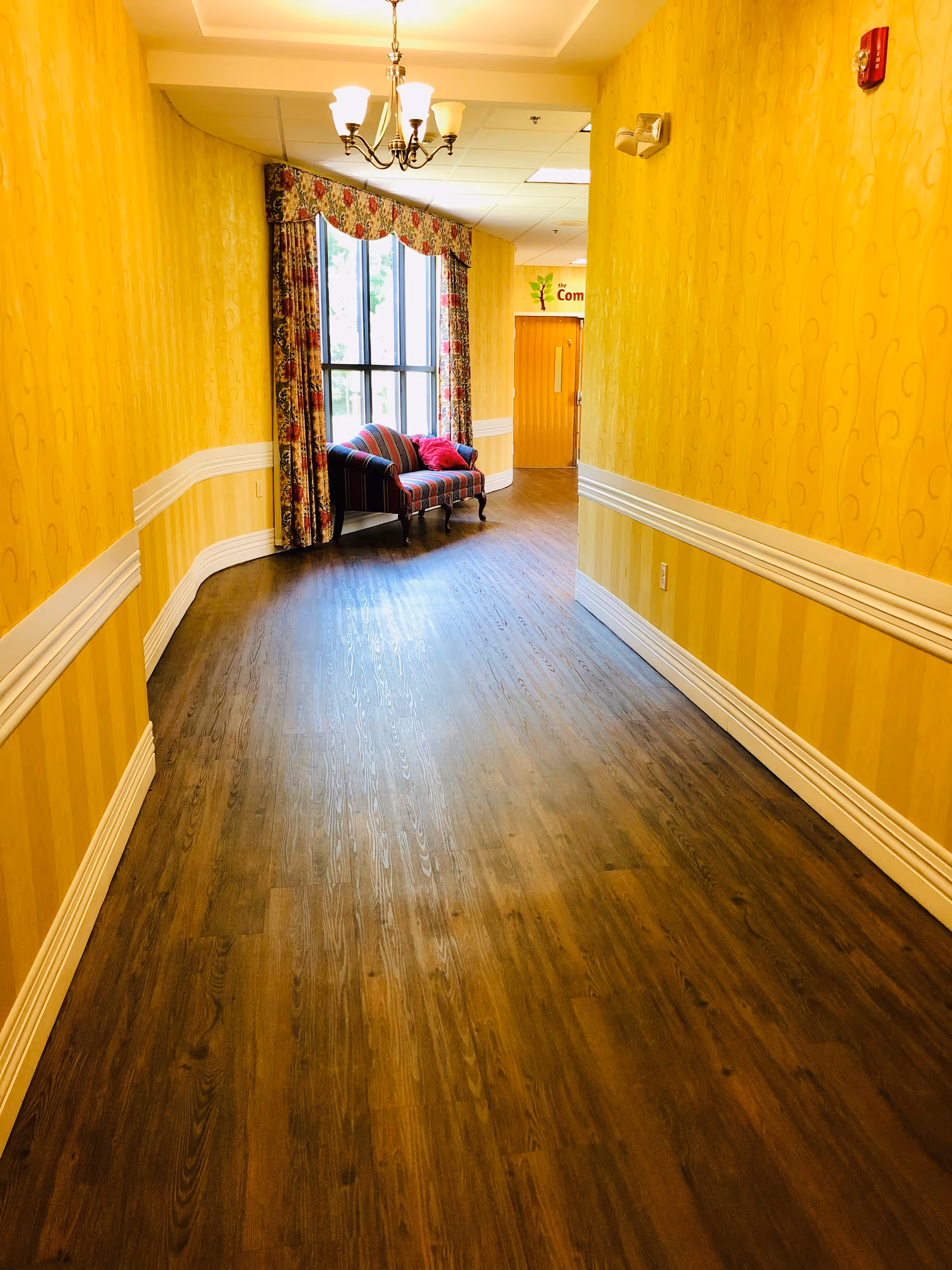 A brightly lit hallway with yellow patterned wallpaper and white trim. At the end of the hallway, there is a large window with floral curtains and a striped upholstered bench with red pillows. A chandelier with five lights hangs from the ceiling.