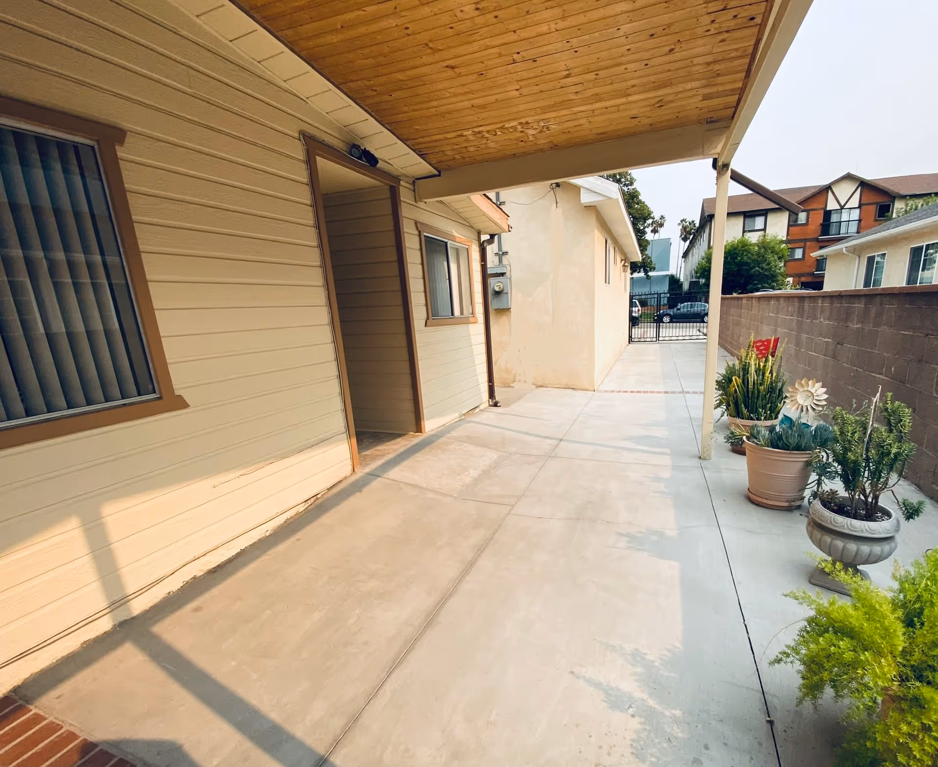 Covered outdoor walkway beside a beige building with windows and a door, concrete floor, several potted plants along a brick wall, and a gated entrance at the end of the walkway.