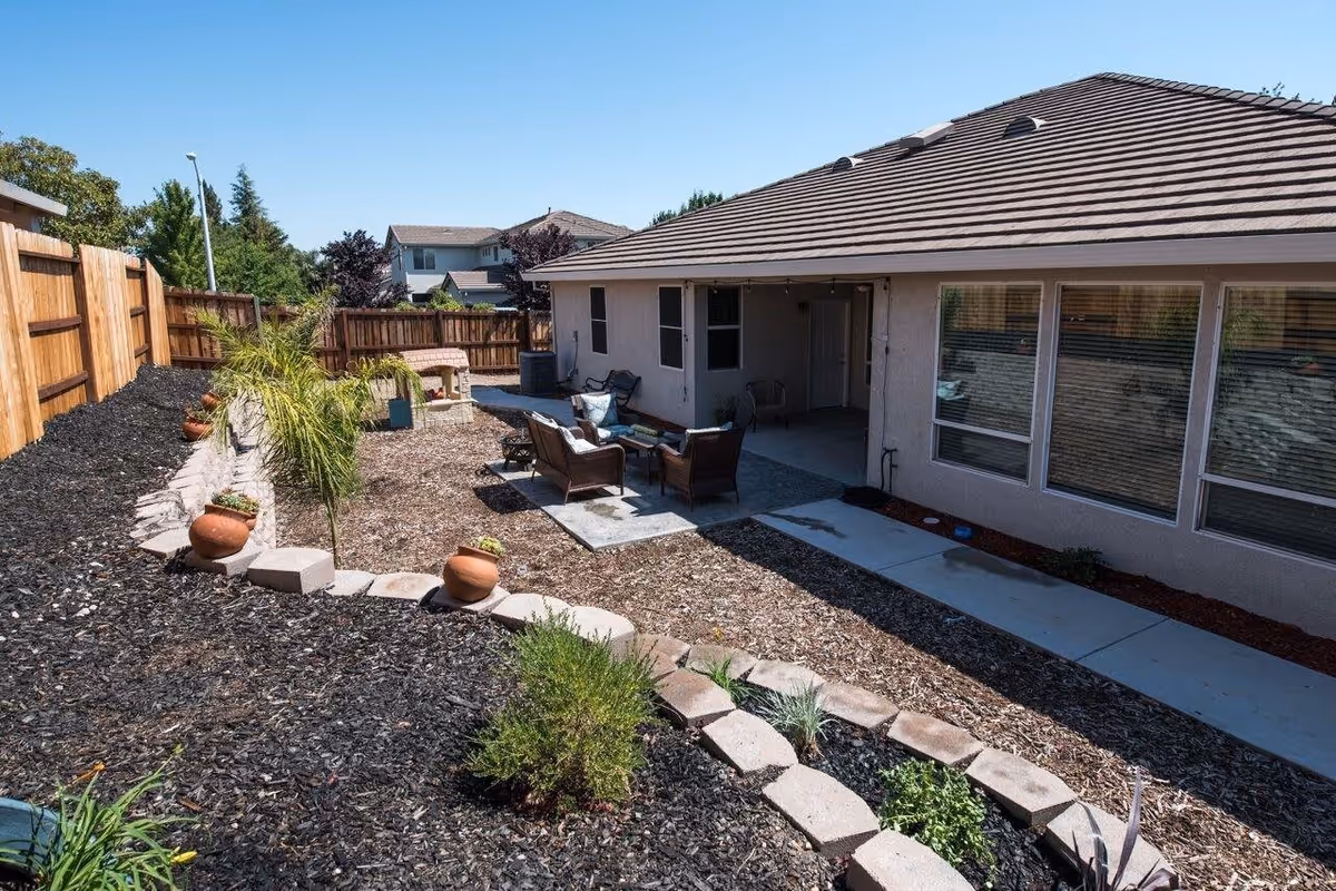 Outdoor patio area of a senior living facility with a seating arrangement including chairs and a small table on a concrete slab. The area is surrounded by a mulched garden with small plants and potted plants along a stone border. The building has large windows and a tiled roof, and a wooden fence encloses the yard under a clear blue sky.
