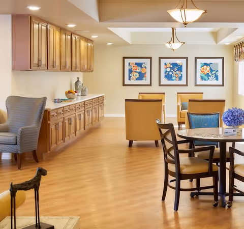 A bright and spacious interior room with wooden flooring and beige walls. There is a long wooden cabinet with multiple drawers and decorative items on top, including a bowl of fruit and vases. The room features several comfortable chairs in yellow and blue upholstery arranged around a round table with a blue flower centerpiece. Three framed floral artworks hang on the far wall. The ceiling has recessed lighting and two hanging light fixtures.