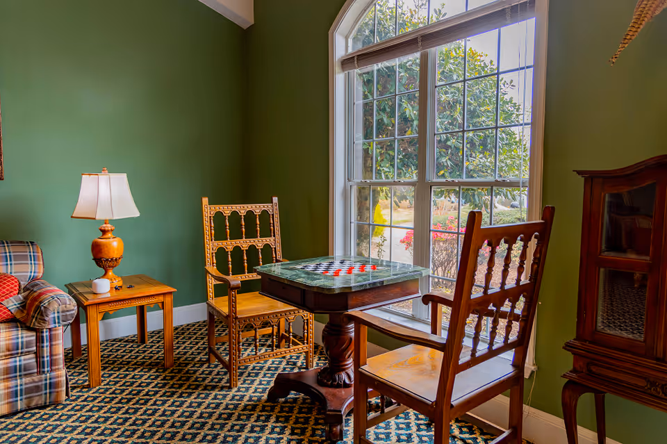 A cozy sitting area with two wooden chairs facing a small table with a checkerboard on it, next to a large window showing greenery outside. The room has green walls, a patterned carpet, a plaid armchair with a red pillow, a wooden side table with a lamp, and a wooden cabinet.