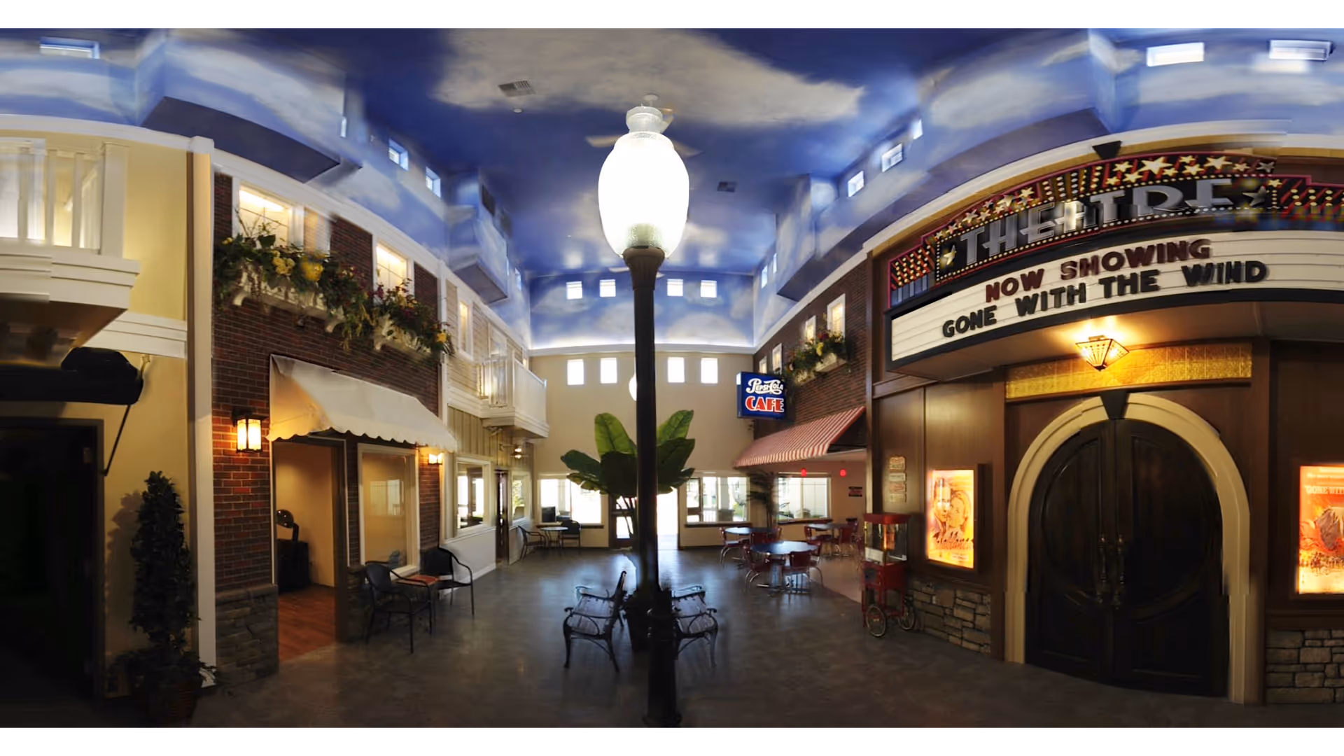 Indoor common area designed to look like a small town street with a sky-painted ceiling, street lamp, benches, storefronts including a cafe and a theater marquee showing 'Now Showing Gone With The Wind'.