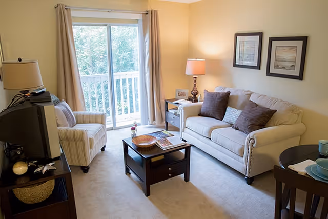 A cozy living room with a beige sofa adorned with three cushions, a striped armchair, a dark wooden coffee table with books and a decorative bowl, a side table with a lamp, and a sliding glass door with beige curtains leading to a balcony with greenery outside.