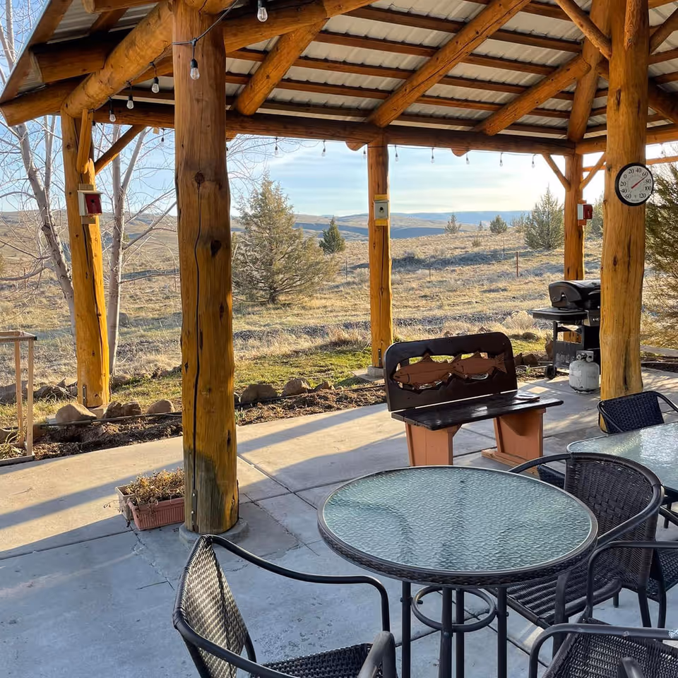 Outdoor covered patio area with wooden beams and a metal roof, featuring a glass-top table with black wicker chairs, a wooden bench, a barbecue grill, and a thermometer on one of the wooden posts. The background shows a natural landscape with trees and hills under a clear sky.