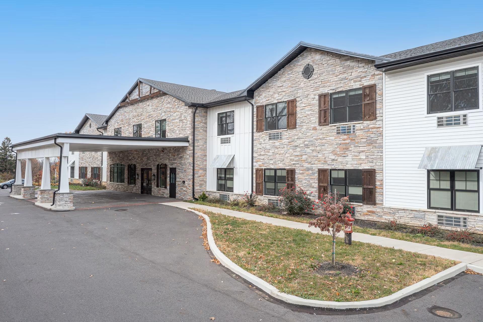 Exterior view of a senior living facility named Atrium of Allentown, featuring a two-story building with stone and white siding, multiple windows with wooden shutters, a covered entrance driveway, and a small landscaped area with grass and a young tree.