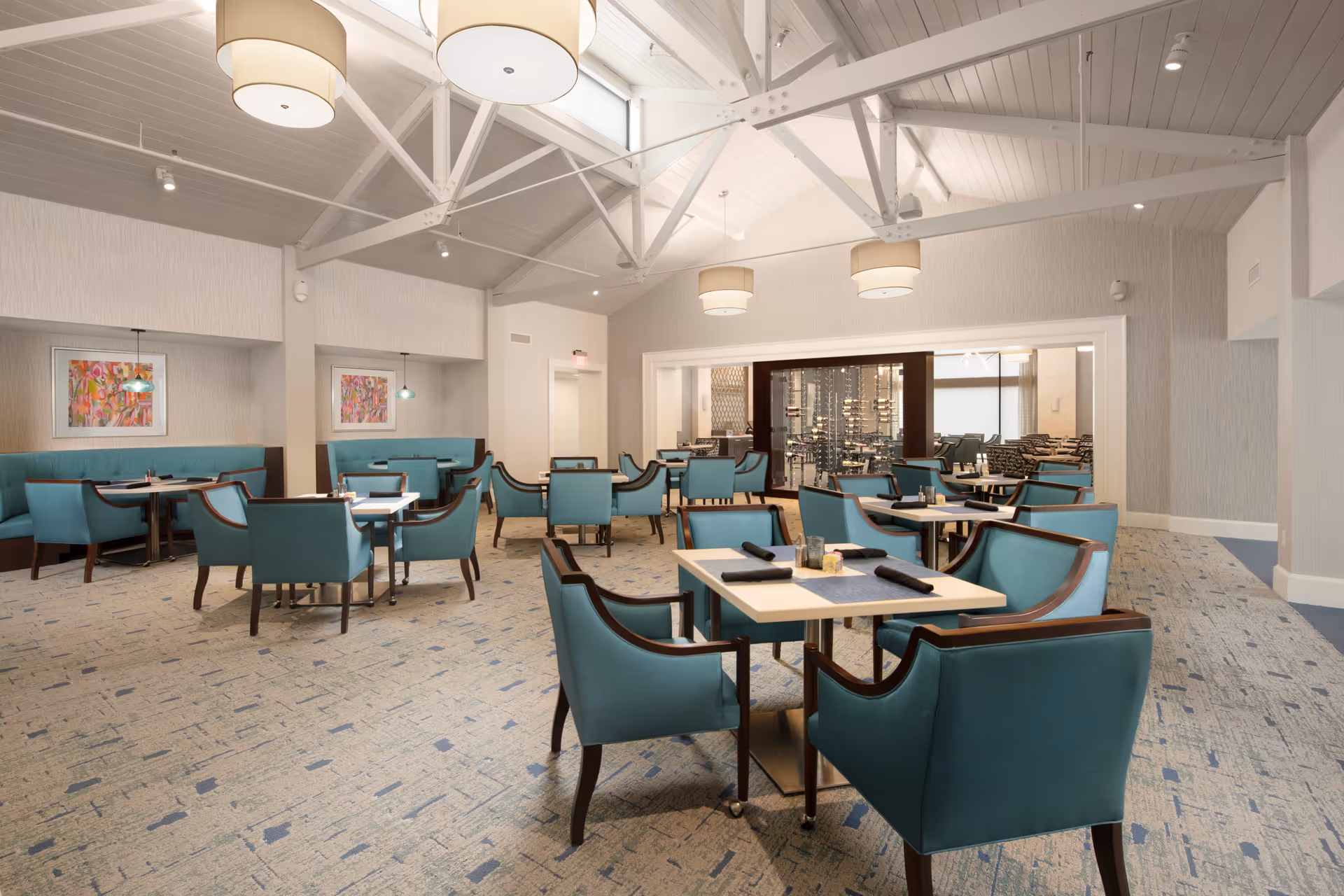 A spacious dining room with multiple tables and teal upholstered chairs arranged neatly. The room features a high vaulted ceiling with exposed white beams and large round pendant lights. There are colorful abstract paintings on the walls and a carpeted floor with a subtle pattern. In the background, a glass-enclosed wine storage area is visible.