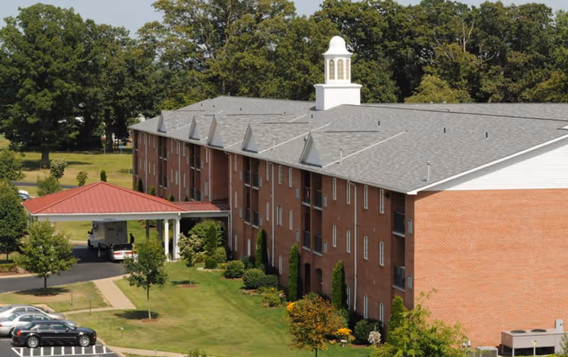 Three-story red-brick apartment building with a covered entrance, parking area, and surrounding trees.