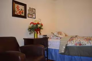 A bedroom in a senior facility with a bed, nightstand, flowers, and wall art.