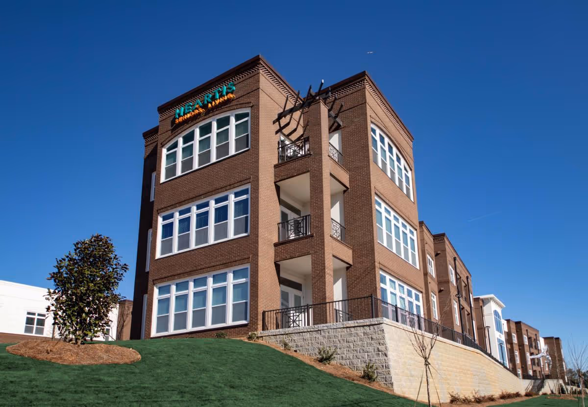 Brick three-story senior living building with large windows, balconies, and a 'Heartis Suwanee Senior Living' sign against a clear blue sky.