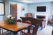 A senior living center common area with a wooden table and chairs in the foreground, a floral centerpiece on the table, and a seating area with a sofa, armchairs, a fireplace, and a wall-mounted TV in the background.