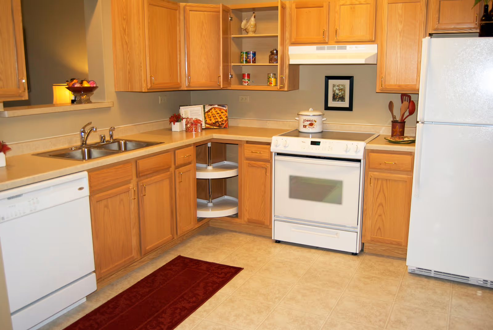 Bright kitchen with oak cabinets, white appliances (stove, refrigerator, dishwasher), double sink, and a red runner rug on tiled floor.