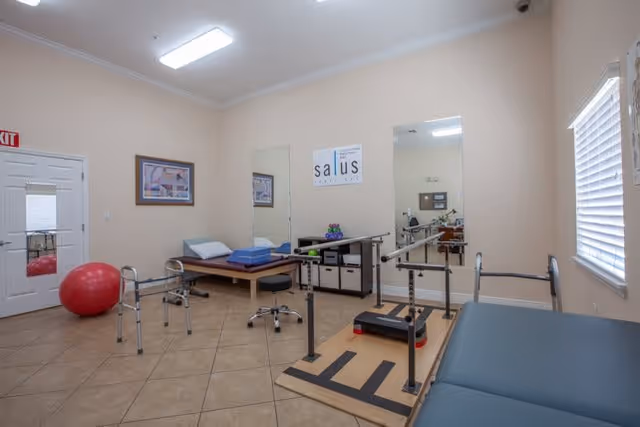A physical therapy or rehabilitation room with exercise equipment including parallel bars, a walker, a red exercise ball, a padded treatment table, and a stool. The room has beige walls, tiled floor, a window with blinds, two large mirrors, and a sign on the wall that reads 'salus'.