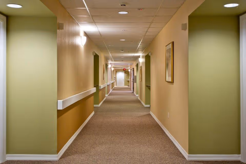 A long, well-lit hallway in a retirement community with beige and green walls, carpeted floor, ceiling lights, handrails along the walls, and framed artwork hanging on the right side.