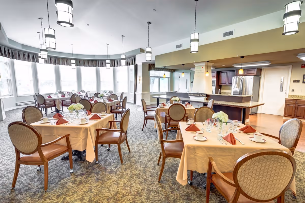 A bright dining room with multiple round tables covered with beige tablecloths, each set with cups, glasses, silverware, and folded red napkins. The room has large windows allowing natural light to fill the space, and there are several pendant lights hanging from the ceiling. The background shows a kitchen area with stainless steel appliances and wooden cabinetry.
